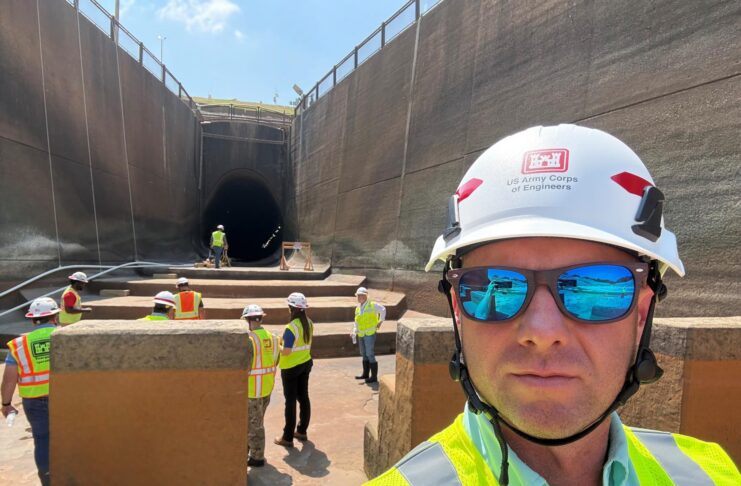 A group of workers in safety vests and helmets stand near a large tunnel under construction; one man is in the foreground.