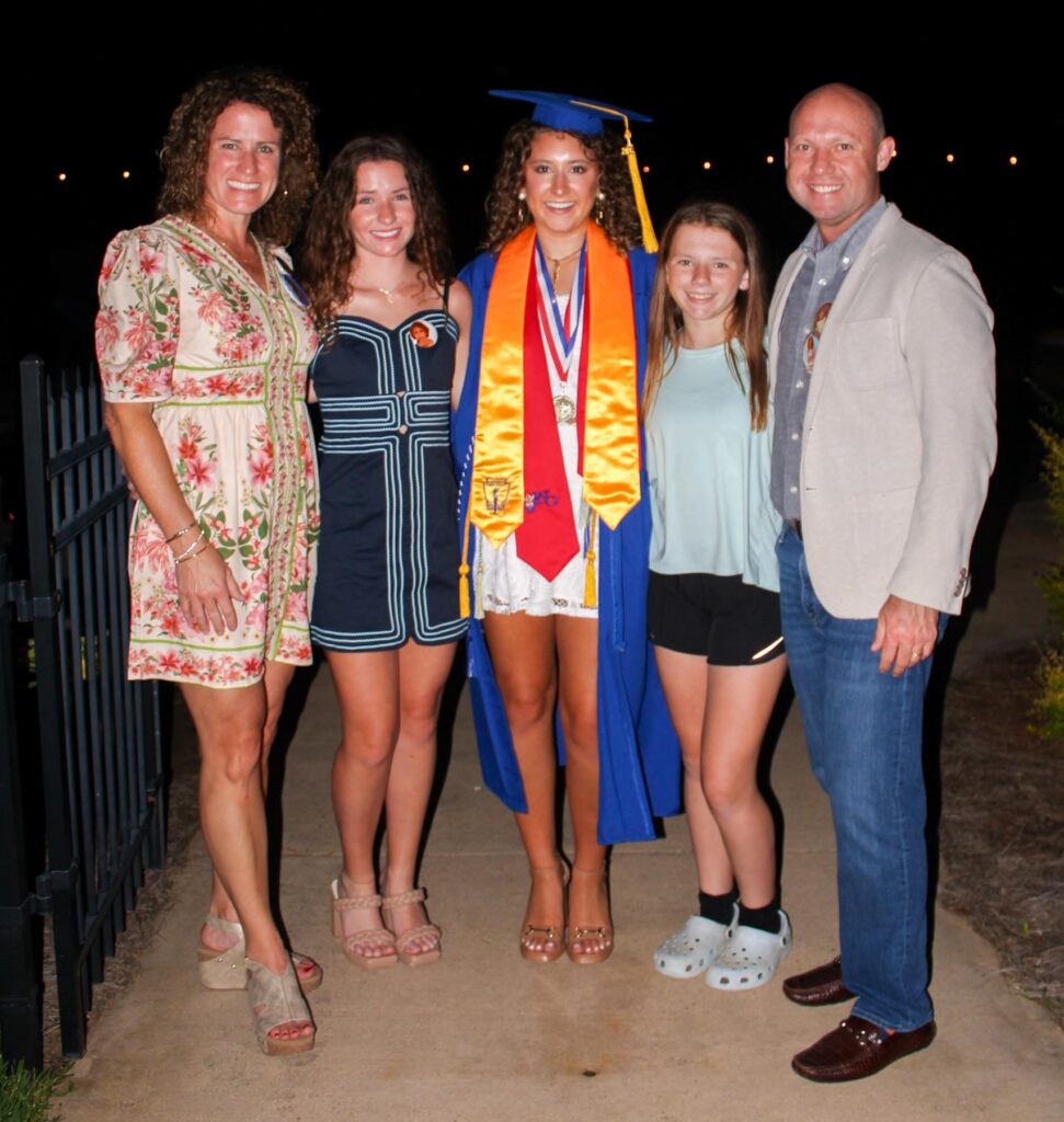 Five people stand together outdoors at night, including one in a graduation cap, gown, and medals.