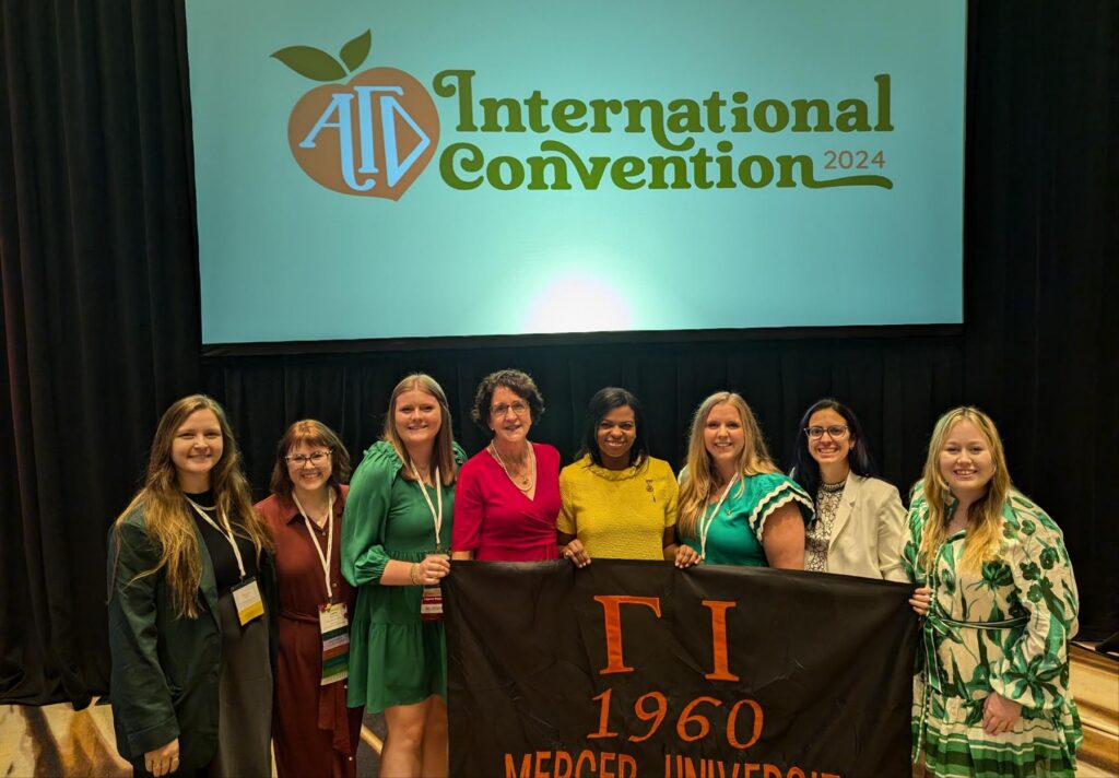 Eight women stand together holding a Mercer University banner at the 2024 International Convention.
