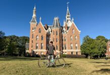 Engineering student develops test rig for bike manufacturer Person standing on grass with a bicycle in front of a large, red-brick building with pointed towers under a clear sky.