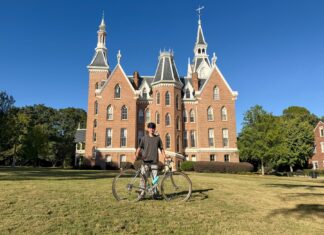Engineering student develops test rig for bike manufacturer Person standing on grass with a bicycle in front of a large, red-brick building with pointed towers under a clear sky.