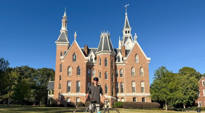 Engineering student develops test rig for bike manufacturer Person standing on grass with a bicycle in front of a large, red-brick building with pointed towers under a clear sky.