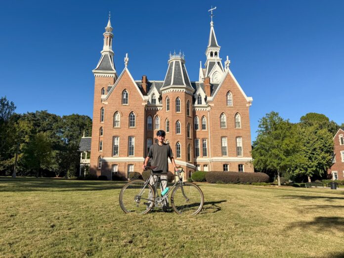 Person standing on grass with a bicycle in front of a large, red-brick building with pointed towers under a clear sky.