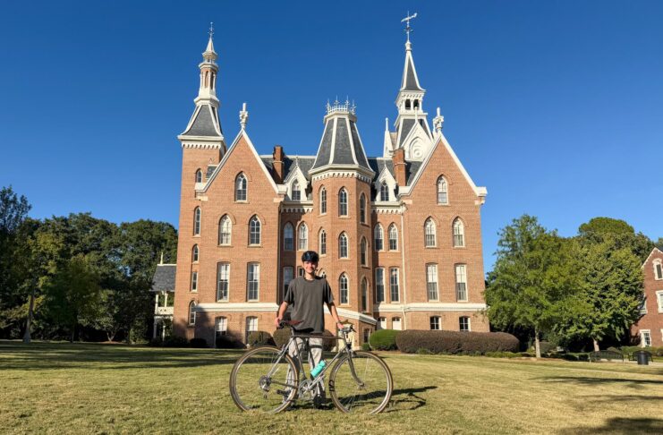 Person standing on grass with a bicycle in front of a large, red-brick building with pointed towers under a clear sky.