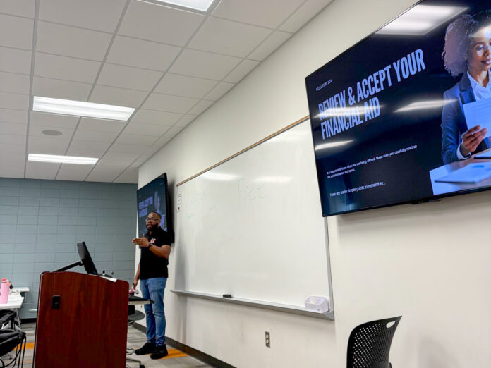 A person stands at a podium in a classroom with a screen displaying Review and Accept Your Financial Aid.