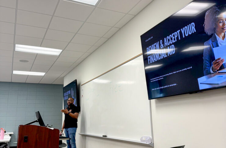 A person stands at a podium in a classroom with a screen displaying Review and Accept Your Financial Aid.