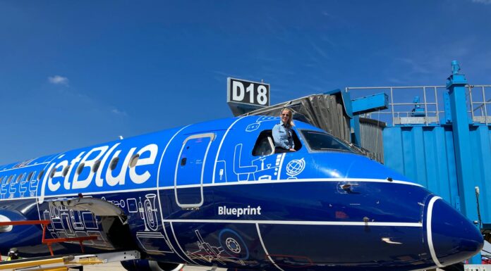Mercer alumna lands high-flying career with JetBlue A pilot leans out of the cockpit window of a parked JetBlue plane with blue livery near gate D18 at an airport.