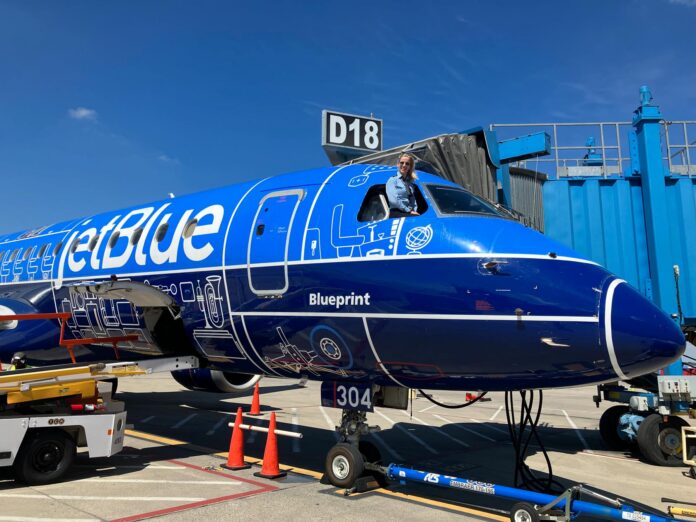 cassie 1 A pilot leans out of the cockpit window of a parked JetBlue plane with blue livery near gate D18 at an airport.