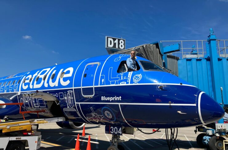 A pilot leans out of the cockpit window of a parked JetBlue plane with blue livery near gate D18 at an airport.
