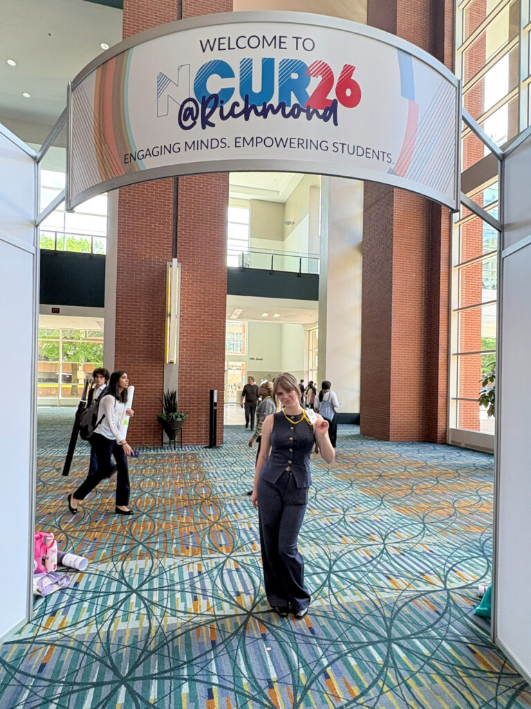 Addie White stands under a CUR 26 @ Richmond welcome sign at a conference center with people in the background.