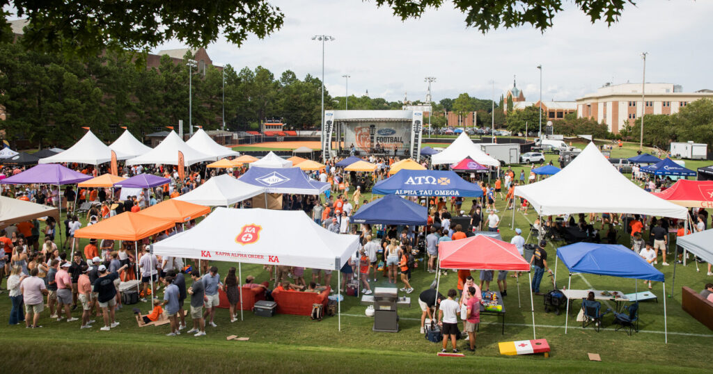 Crowd gathers under colorful tents at an outdoor event with a stage, trees, and campus buildings in the background.