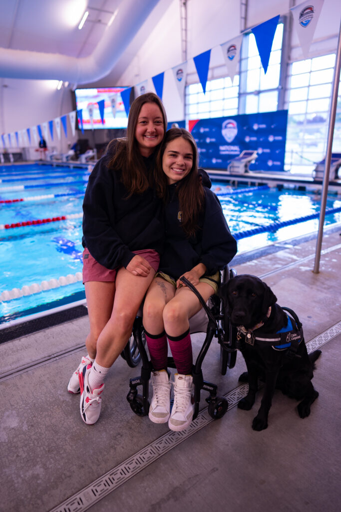 Two young women, one in a wheelchair, sit by an indoor pool with a black service dog beside them.