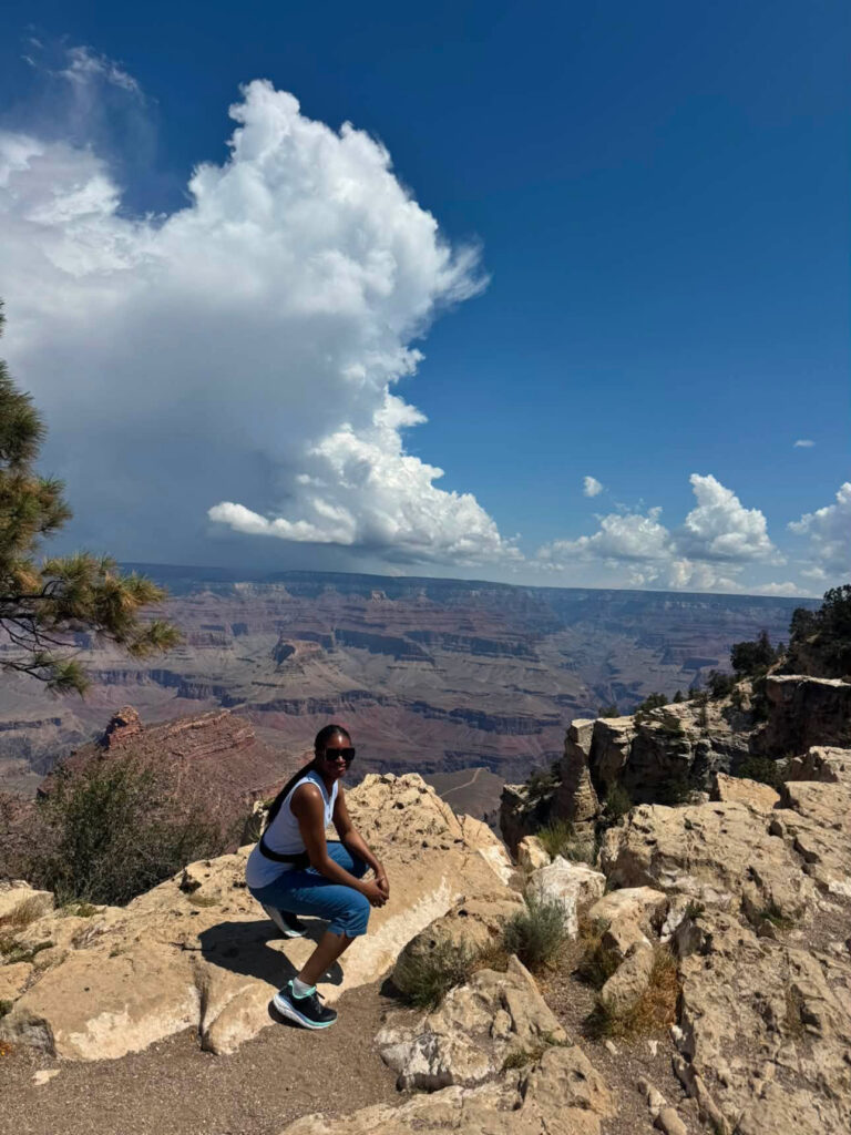 Susanna Nwuha squats on a rocky ledge overlooking the Grand Canyon under a partly cloudy sky.