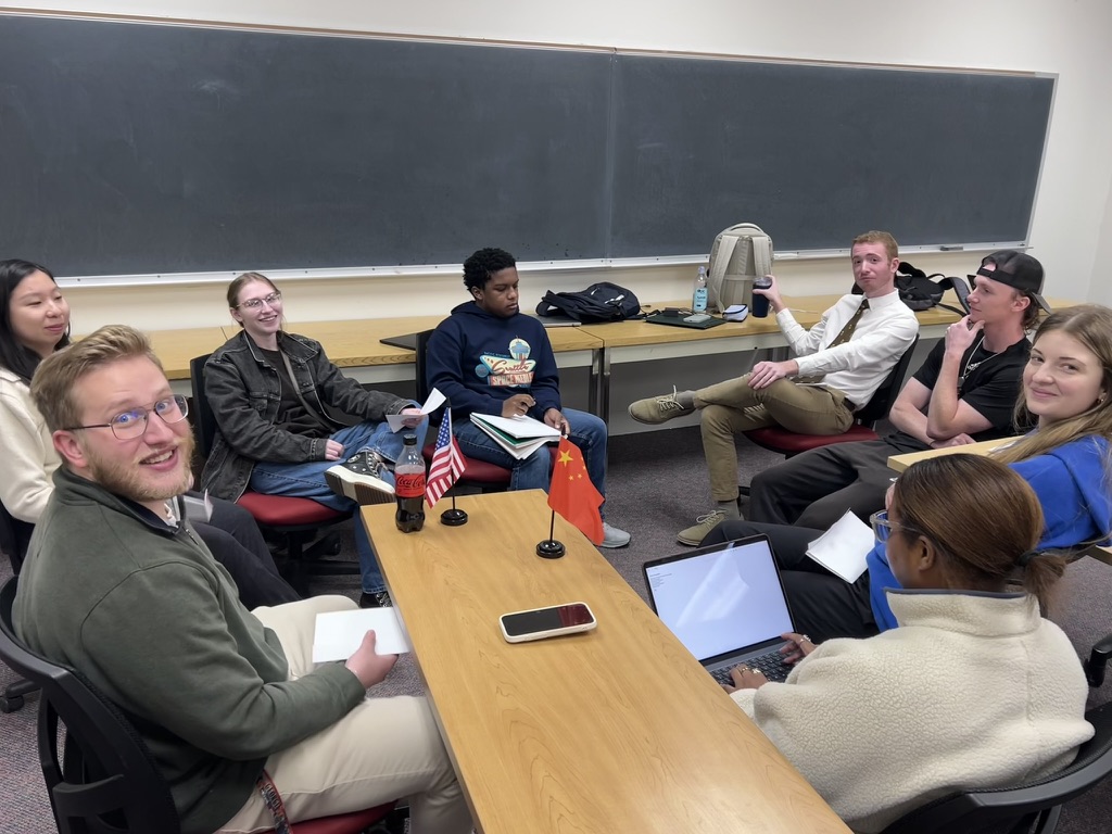 Eight people sit around a table with notebooks and laptops, two small flags at the center, in a classroom setting.