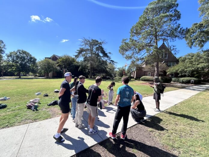 A group of people stands and talks on a sidewalk next to a grassy area on a sunny day.