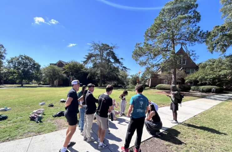 A group of people stands and talks on a sidewalk next to a grassy area on a sunny day.