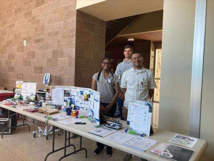 Three people stand behind a table displaying healthcare pamphlets and medical equipment in a well-lit hallway.