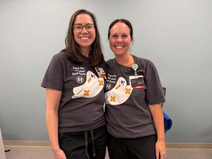 rebekah carter 1 Two women wearing matching shirts with cartoon ghosts stand side by side, smiling in a room with a light blue wall.