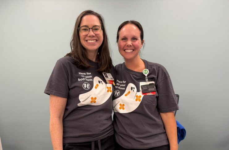 Two women wearing matching shirts with cartoon ghosts stand side by side, smiling in a room with a light blue wall.