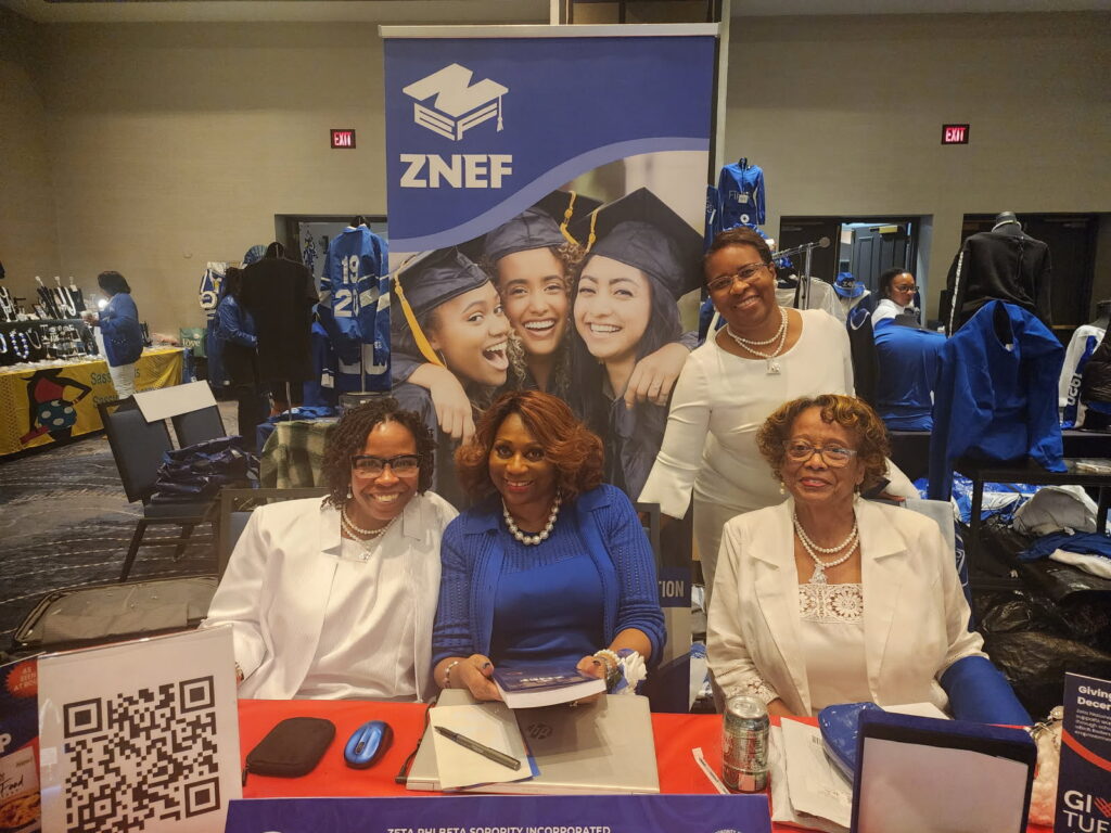 Four women sit at a ZNEF booth with promotional materials; a banner with graduates is displayed behind them.