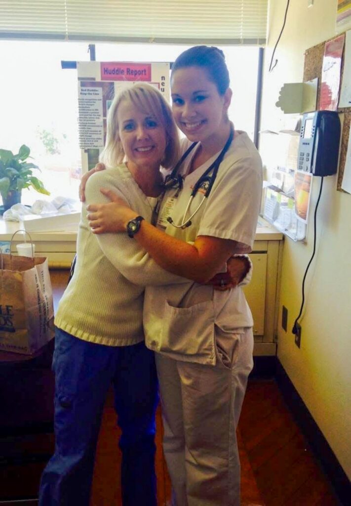 Two women, one in scrubs and one in casual clothes, stand hugging in a brightly lit room with medical equipment.