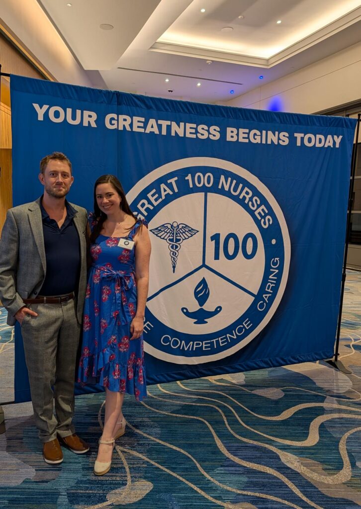 Two people stand in front of a Great 100 Nurses banner at an indoor event.
