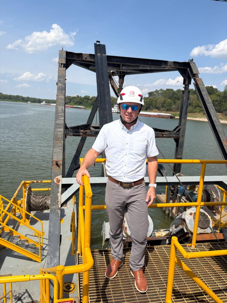 Man in hard hat and sunglasses stands on industrial platform by water, with trees and blue sky in background.