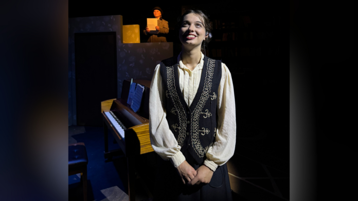 Young woman in vintage clothing stands in front of a piano on stage; another person reads from paper in the background.