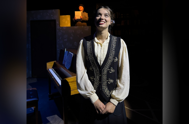 Young woman in vintage clothing stands in front of a piano on stage; another person reads from paper in the background.