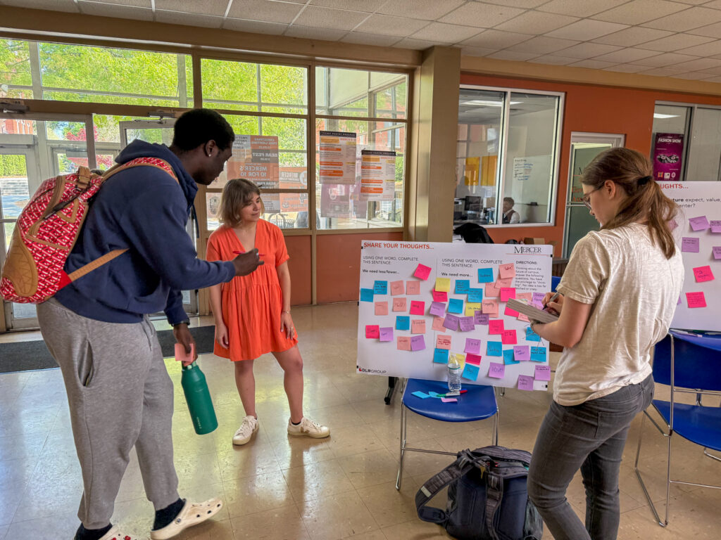 Three Mercer students stand by a board covered in colorful sticky notes, discussing and adding more notes.