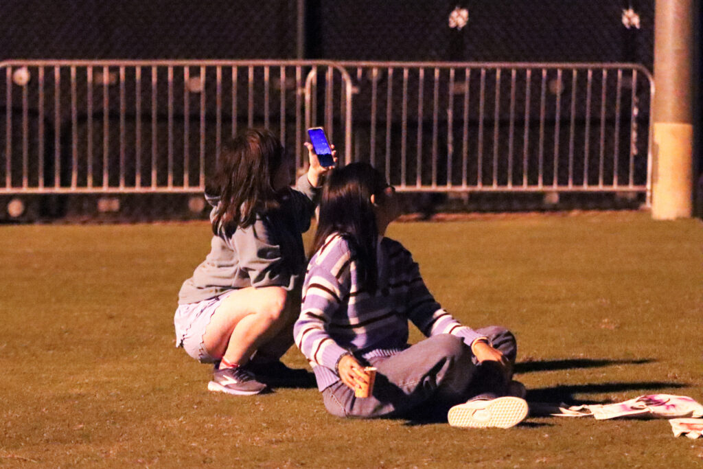 Two people sitting on the grass at night, one holding up a smartphone to record or take a photo.