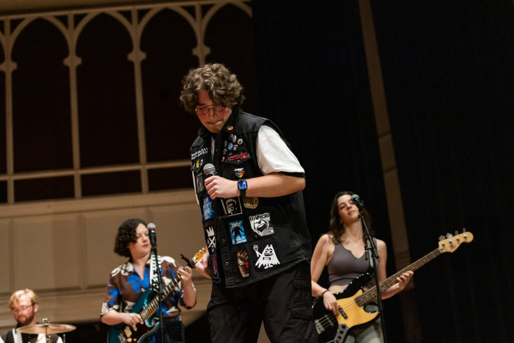 A band performing on stage, with a lead singer in a patch-covered vest, a guitarist, and a bassist.