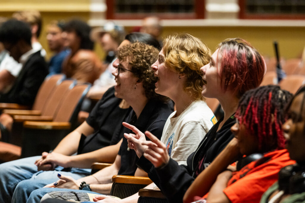 Audience members sits in a theater, laughing and pointing toward a stage.