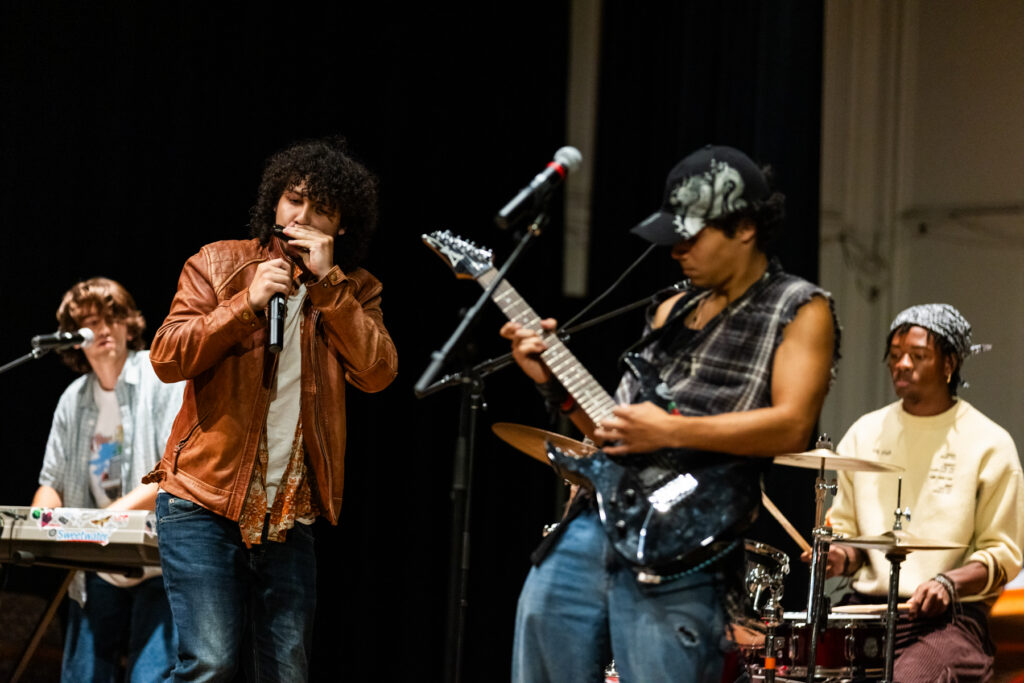 A band performing on stage, featuring a harmonica player, a guitarist, a drummer, and a keyboardist.