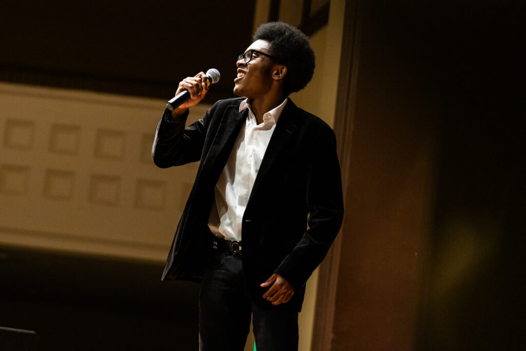 A student in a blazer and white shirt sings into a microphone on stage.
