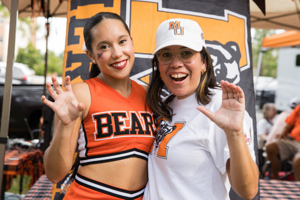 A cheerleader in an orange uniform and a woman in a white cap and t-shirt pose with their hands raised in front of a banner.