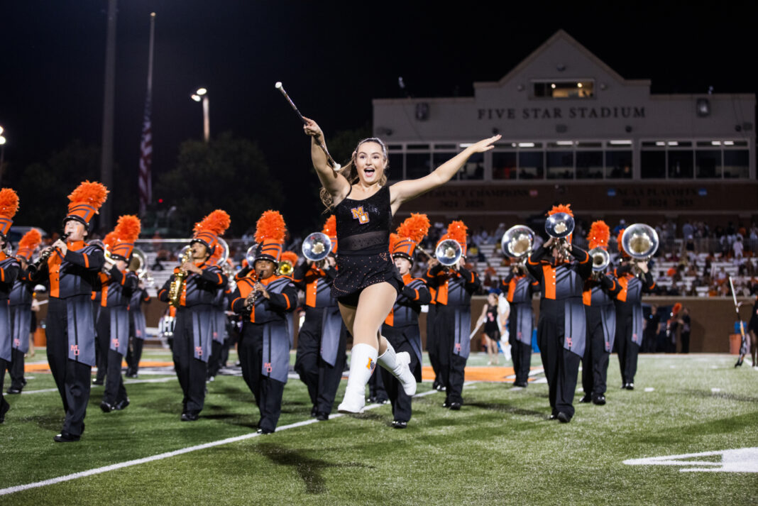 A baton twirler leaps in front of a marching band on a football field at night.