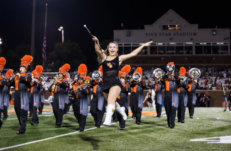 A baton twirler leaps in front of a marching band on a football field at night.