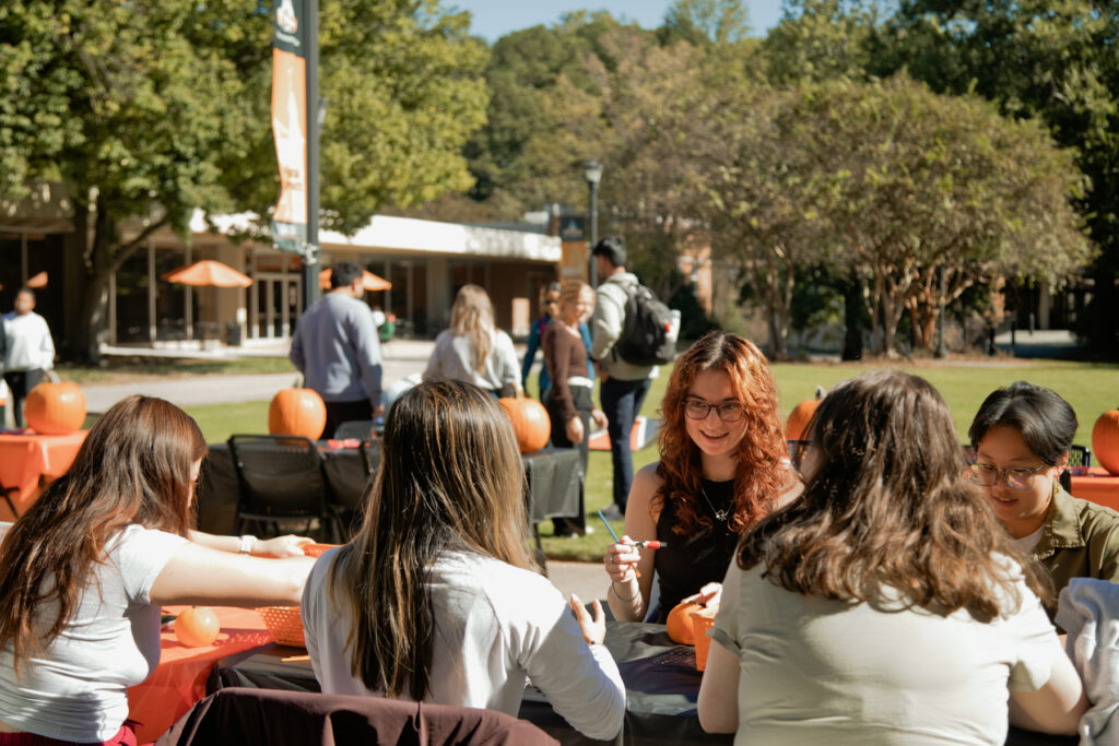 A group of students sitting at a table outdoors painting small pumpkins.