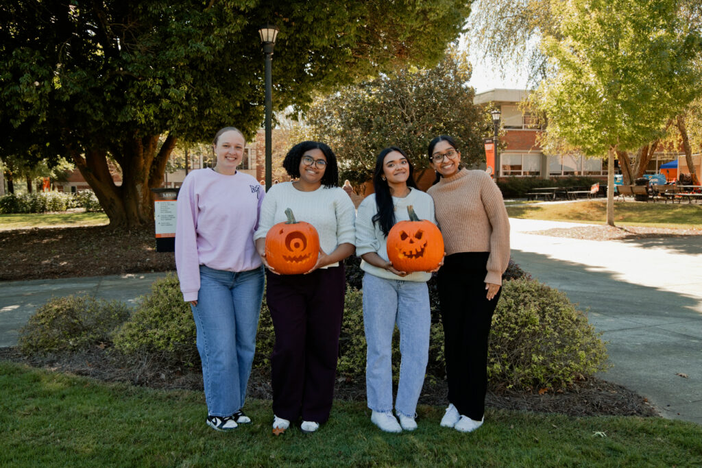 Four young women stand outdoors, two of them holding carved pumpkins.