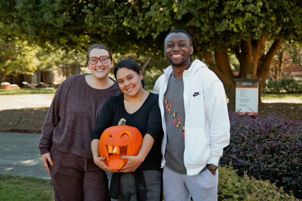 Three students smiling outdoors, one holding a carved pumpkin.