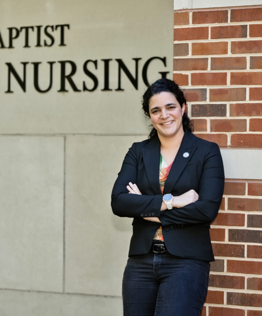 A woman in a black blazer stands with arms crossed in front of a building wall labeled Baptist Nursing.