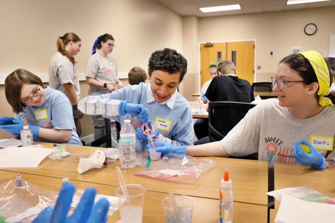 Students wearing blue gloves conduct a science experiment with plastic cups and liquids at a table.