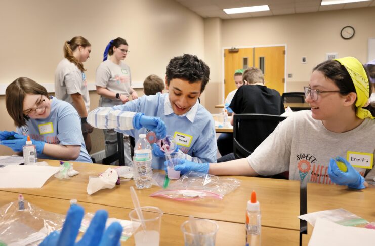 Students wearing blue gloves conduct a science experiment with plastic cups and liquids at a table.