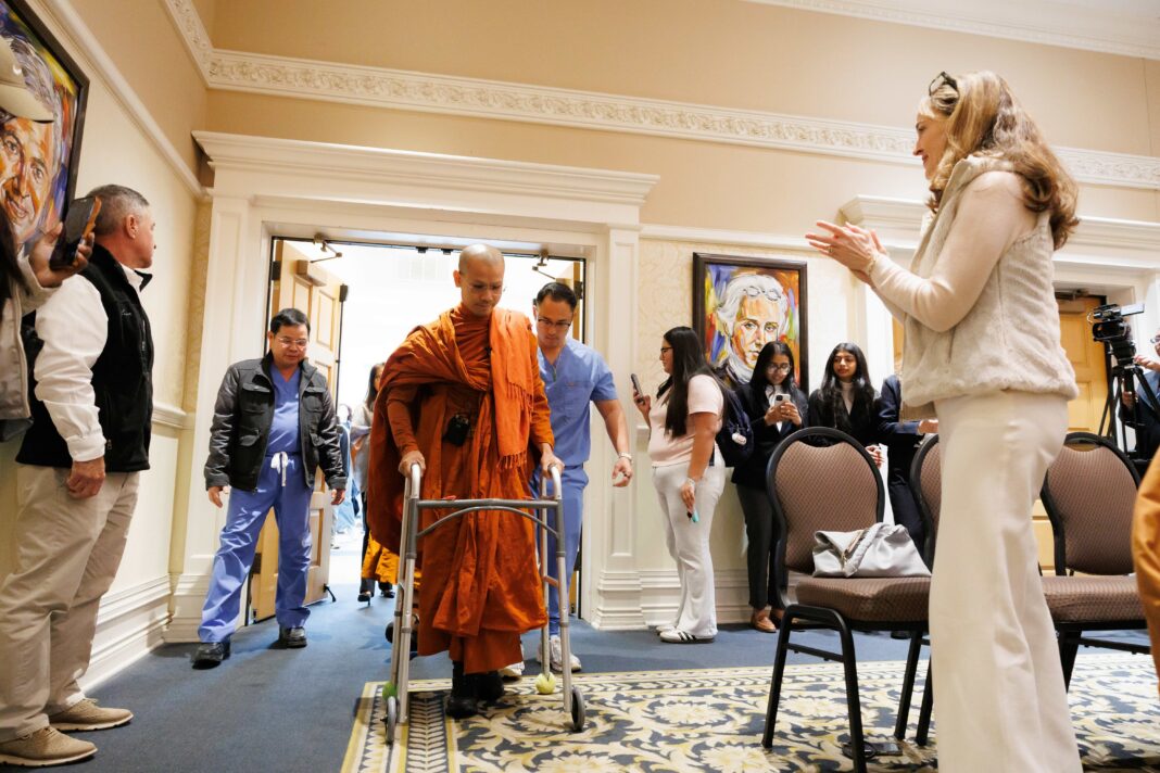 260318MUMonkProsthetic_0444 A monk in orange robes walks with a walker through a doorway as people watch and record with phones.