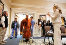 Buddhist Monk fitted with prosthetic by Mercer On Mission team to lead Macon Community Peace Walk A monk in orange robes walks with a walker through a doorway as people watch and record with phones.