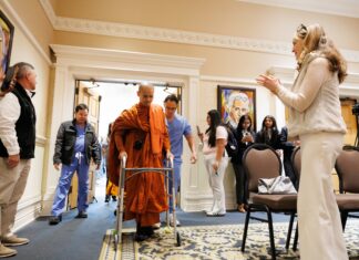 Buddhist Monk fitted with prosthetic by Mercer On Mission team to lead Macon Community Peace Walk A monk in orange robes walks with a walker through a doorway as people watch and record with phones.