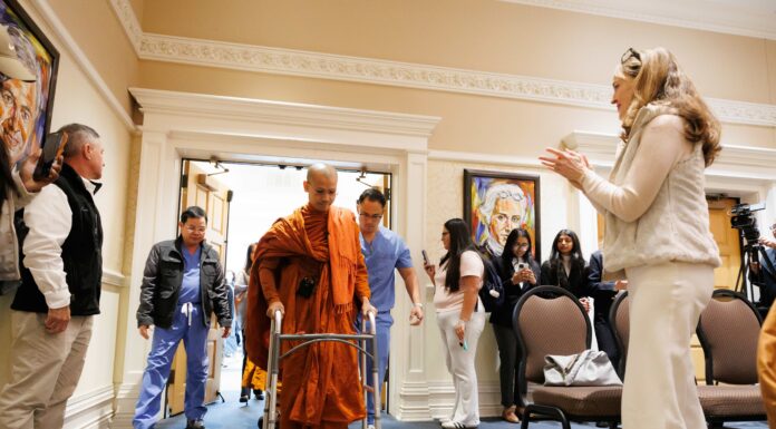 Buddhist monk fitted with prosthetic by Mercer On Mission team to lead Macon Community Peace Walk A monk in orange robes walks with a walker through a doorway as people watch and record with phones.