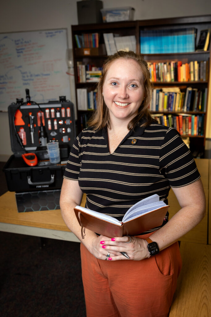 A woman in a striped polo shirt holds an open notebook, with a technical equipment case and a bookshelf in the background.