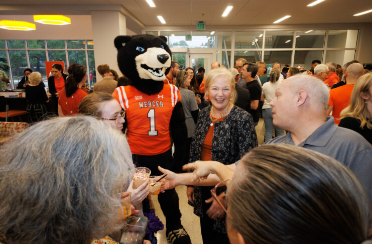 A Mercer University bear mascot stands in a crowded room with people, including the Mercer president, holding small ice cream cups.